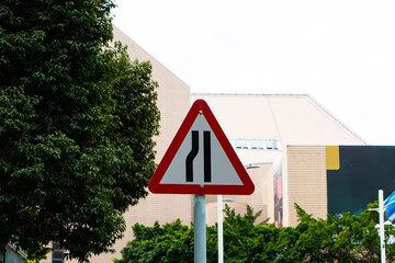 Triangle road sign aluminum warning left narrow on road with trees and building with bright sunlight is background. Traffic sign symbols to warn passing vehicles to be careful in driving area
