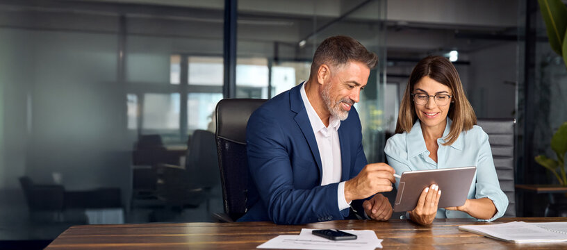 Two colleagues, group of partners, professional people working together. Mature 50s age Latin business man mentoring mid age European woman discussing project on tablet in office. Banner, copy space