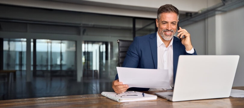 Smiling businessman talking phone, check database in office. Latin or Indian male business man holding documents, working at laptop computer doing online trade market tech research. Banner, copy space - Powered by Adobe