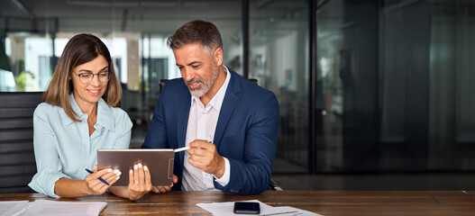 Mature 50s age Latin business man mentoring mid age European woman discussing project on tablet in...