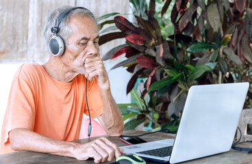 Senior man with grey hair wearing headphones and an orange shirt working on a laptop outdoors on a wooden table, touching his chin in concentration.