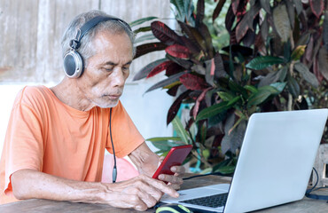 Elderly man using laptop outdoors with headphones, surrounded by lush greenery. Digital connection in a rural setting.