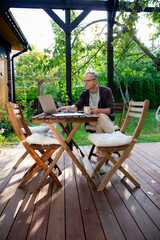 Middle aged man working remotely on laptop at cozy sunny patio, digital nomad lifestyle, outdoor home office, freelance and productivity concept