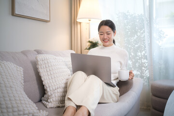 Asian woman sitting on a couch with a laptop and coffee mug, smiling while working from home.