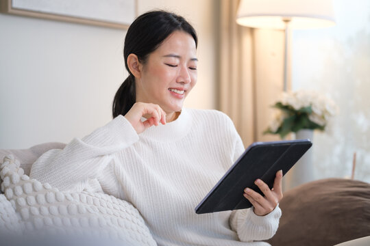 Young woman in cozy white sweater using digital tablet in bright modern living room.