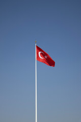 Red Turkish flag with crescent and star waving on a tall flagpole against a clean blue sky. Perfect for stories on Turkey, patriotism, national days, travel, politics, news design, or backgrounds.