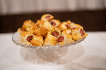 Delicious Golden Brown Savory Pastry Wrapped Appetizers Filled with Mini Sausages Arranged on a Clear Glass Pedestal Dish for a Festive Gathering