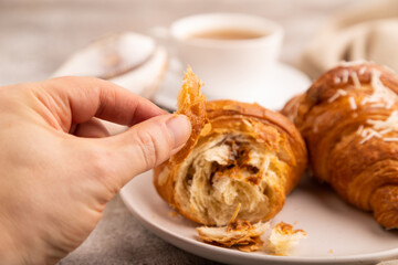 Croissant on white plate with hand on brown concrete background, cup of coffee, side view, close up, selective focus