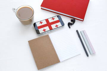 Smartphone with the flag of Georgia on screen lying on a white desk with notebook, pen, coffee, and headphones. Concept of learning Georgian language, education, travel, and modern study tools