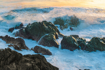 Fototapeta premium Dynamic coastal action, foaming wave collides with rocky headland under warm golden light.