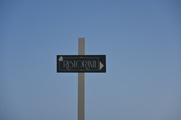 A restaurant sign (italian: Ristorante) with an arrow pointing direction, set against a clear blue sky background