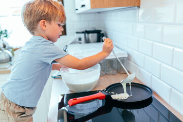 Child about 5 years old pouring pancake batter on pan with ladle in kitchen