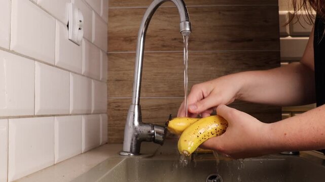 Person washes bananas under running water in kitchen. Water flows from faucet and cleans banana peel. Banana washing shows food hygiene and healthy lifestyle.