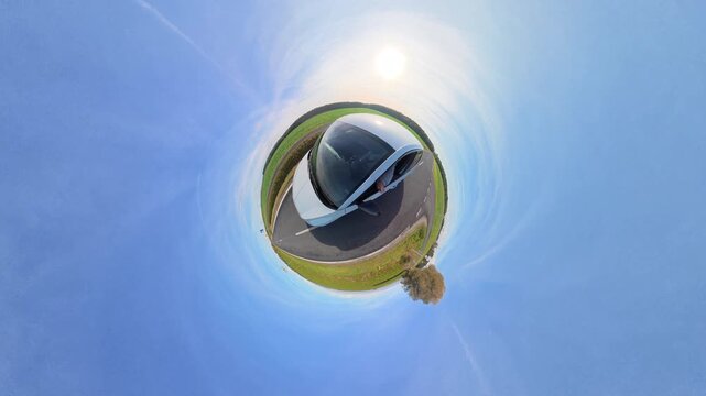 A car travels on a road, surrounded by lush fields under a bright sky. The panoramic lens gives a spherical effect, creating a mini-planet impression. 360 view 