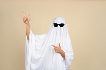 Woman in Halloween ghost costume and sunglasses pointing at something on beige background.