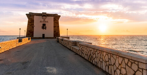 beautiful sunset or sunrise at beautiful embarkment with nice tower on a cape point of a promontory among blue sea vawes. Urban dock pier in golden evening sunset rays leading to a scenic tower.