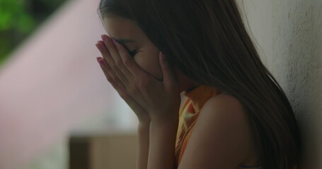 Young girl with brown hair, covering her face with her hands while leaning against a wall outdoors,...