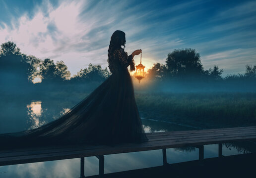 photo real person silhouette gothic woman hand hold lantern lamp walking across bridge over river dense fog mist night nature dark dramatic blue sky lake. long black dress back rear view. Fantasy girl
