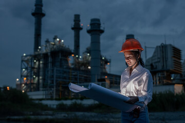Female industrial engineer wearing safety helmet holding blueprints during night shift at power plant, symbolizing leadership, planning, and diversity in engineering and construction industry.