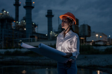 Female industrial engineer wearing safety helmet holding blueprints during night shift at power plant, symbolizing leadership, planning, and diversity in engineering and construction industry.