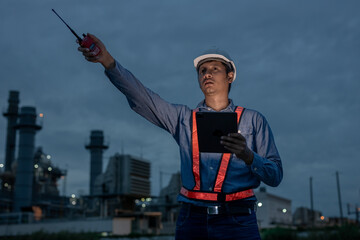 Industrial engineer in safety gear using walkie-talkie and tablet while giving direction during night shift at refinery or power plant, focusing on real-time communication and coordination.