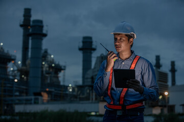 Industrial engineer in safety uniform using walkie-talkie and digital tablet during night shift inspection at oil refinery or power plant, ensuring system safety and operations.