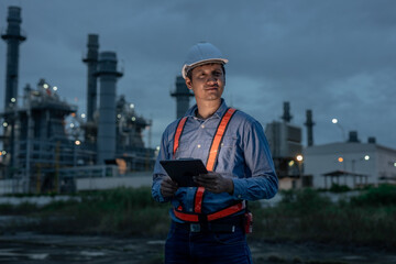 Industrial engineer in safety gear using digital tablet during night shift at power plant or refinery, monitoring operational data and ensuring accuracy in industrial facility management.