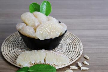 Pomelo flesh is arranged overflowing in a black ceramic bowl, decorated with bright green leaves, and placed on a round base on a wooden table.

