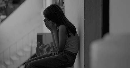 Young child covering face with hands, eyes closed, sitting on the floor in an emotional moment,...