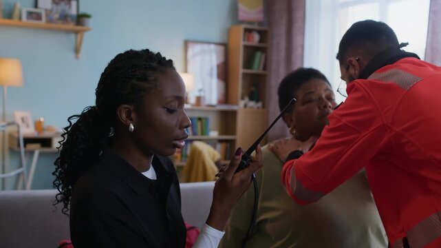 Handheld shot of female medical worker using radio while male paramedic in uniform evaluating health condition of elderly woman at home