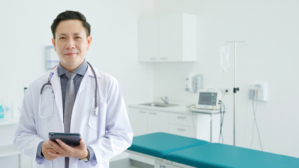 Portrait of Asian senior doctor standing while using tablet in Examination Room. COPY SPCAE.