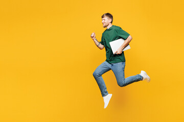 Obraz premium Full body side view happy young man he wear green t-shirt casual clothes jump high hold in hand closed laptop pc computer isolated on plain yellow orange background studio portrait. Lifestyle concept.