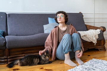 Calm millennial woman feels autumn blues sits on wooden floor at home with cat, gently stroking pet deep in thoughts. Serene young female resting after busy day petting beloved cat, melancholy mood.