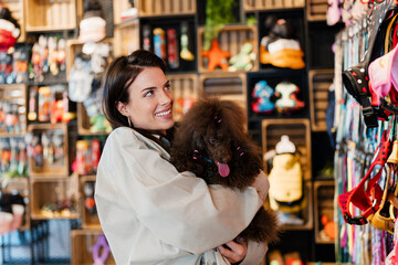 Woman smiling and holding her poodle dog while looking at colorful pet harnesses and supplies in a retail pet shop, conceptualizing pet ownership and care