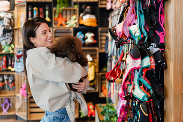 Woman smiling and holding her poodle dog while looking at colorful pet harnesses and supplies in a retail pet shop, conceptualizing pet ownership and care