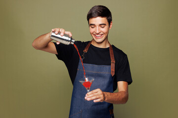Young man barista bartender barman employee wearing jeans apron t-shirt work in night club hold shaker make alcohol martini cocktail isolated on plain green background. Small business startup concept.