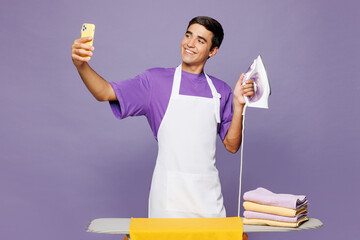 Young man wears violet t-shirt iron clothes on ironing board while doing housework tidy up do selfie shot on mobile cell phone isolated on plain pastel light purple background. Housekeeping concept.