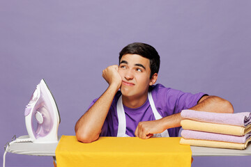 Young minded sad frustrated depressed man he wearing violet t-shirt iron clothes on ironing board while doing housework tidy up isolated on plain pastel light purple background. Housekeeping concept.