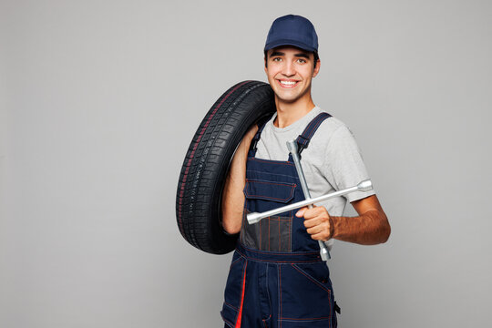 Young smiling happy car mechanic man he wearing overall hat uniform workwear clothes work in garage holding in hand cross wrench tool isolated on plain grey background. Automotive repair job concept.