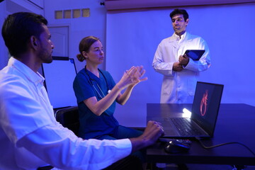 group of doctors is having a treatment planning meeting and a handsome Caucasian male doctor presents at the front of the room, discussing research data with slides on the wall.