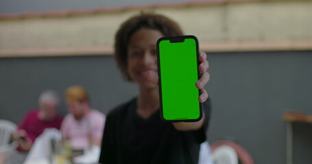 Teenager holding smartphone with green screen, smiling confidently, outdoors at a casual gathering with diverse adults interacting in the blurred background