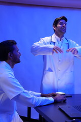 group of doctors is having a treatment planning meeting and a handsome Caucasian male doctor presents at the front of the room, discussing research data with slides on the wall.