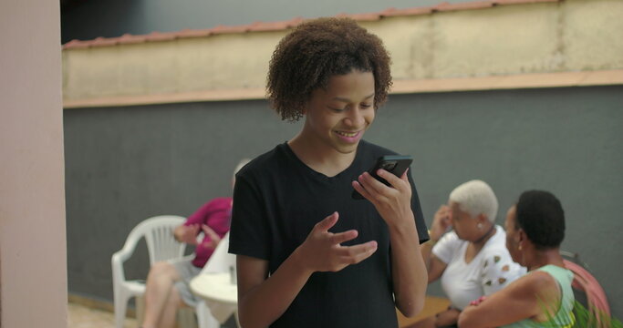 Teenager of African descent showcasing smartphone with green screen, chroma key, outdoors in a social setting with diverse adults enjoying casual conversations in the background