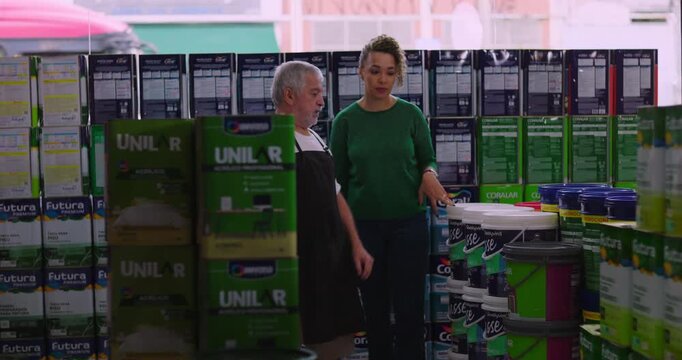 Elderly male shopkeeper interacting with a smiling female customer with curly hair in a hardware store aisle, surrounded by shelves stocked with items