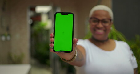 African American woman extending smartphone forward, green screen Chroma Key visible, confident smile, blurred background, emphasizing digital interface and connection outdoors