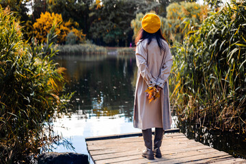 Back view of a girl in a beige coat and yellow hat standing on a wooden pier by the lake.