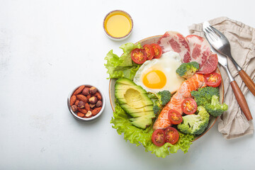 Healthy ketogenic diet concept showing a balanced meal on plate with salmon, avocado, fried egg, bacon, broccoli, tomato, and fresh salad, promoting low carb and high fat eating, copy space