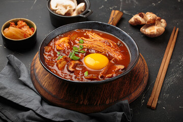Top view of spicy kimchi jjigae soup in a black bowl with egg yolk, enoki, shiitake, tofu and green onions, with chopsticks and cooking ingredients, cuisine of Korea