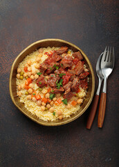 Traditional dish of Maghreb cuisine, a Moroccan couscous with lamb or beef, vegetables, chickpeas, and peas in a rustic bowl with cutlery on dark background