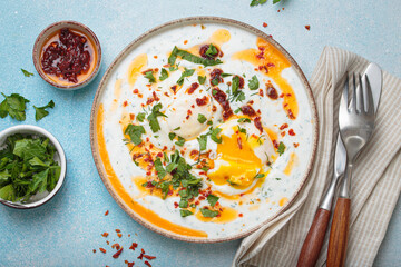 Turkish eggs cilbir, a delicious and colorful breakfast with poached eggs on yogurt, seasoned with spices and fresh herbs, arranged on a blue background